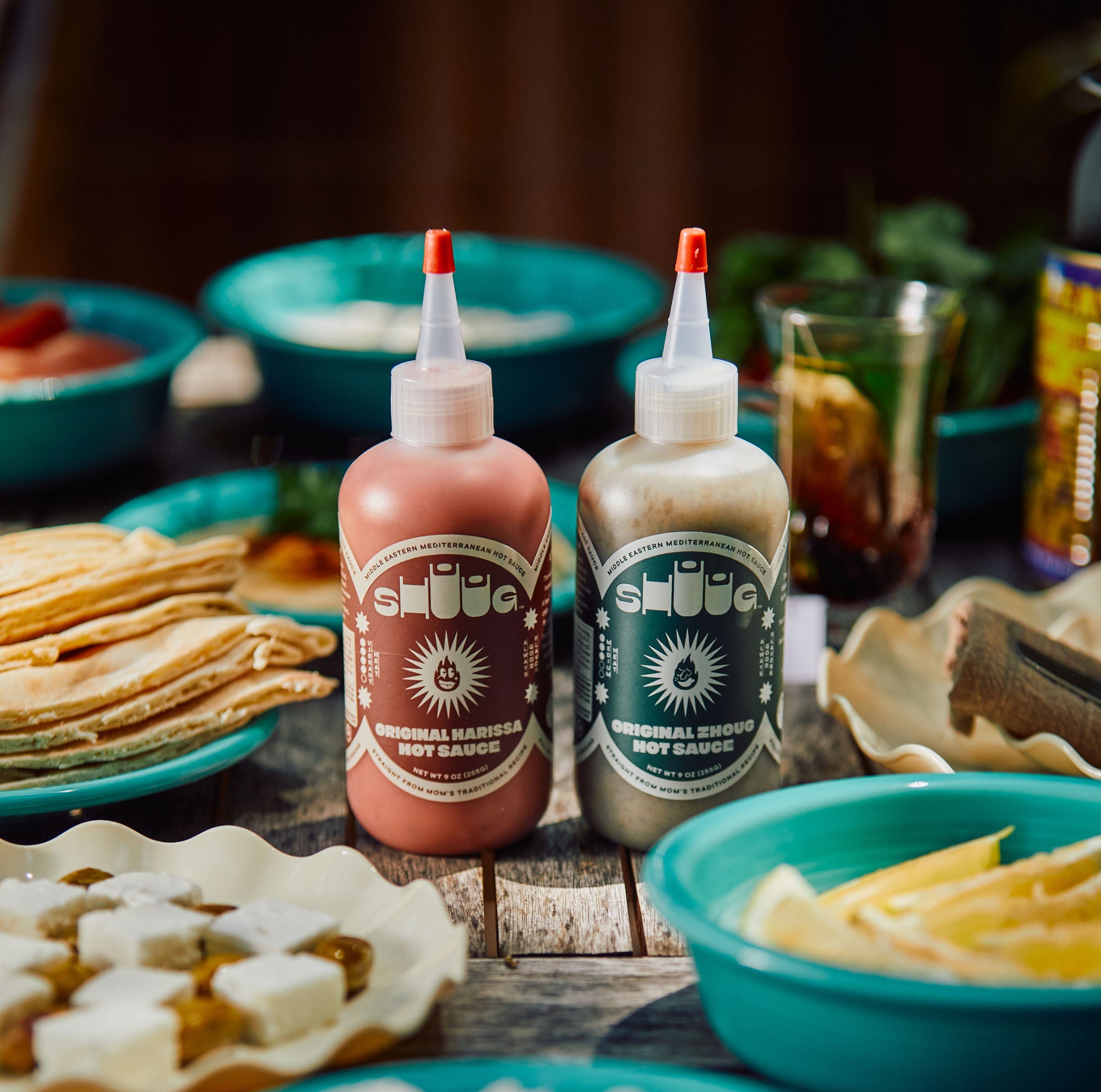 Two Bottles of Shuug Original Zhoug and Harissa Hot Sauce on a Table of Food